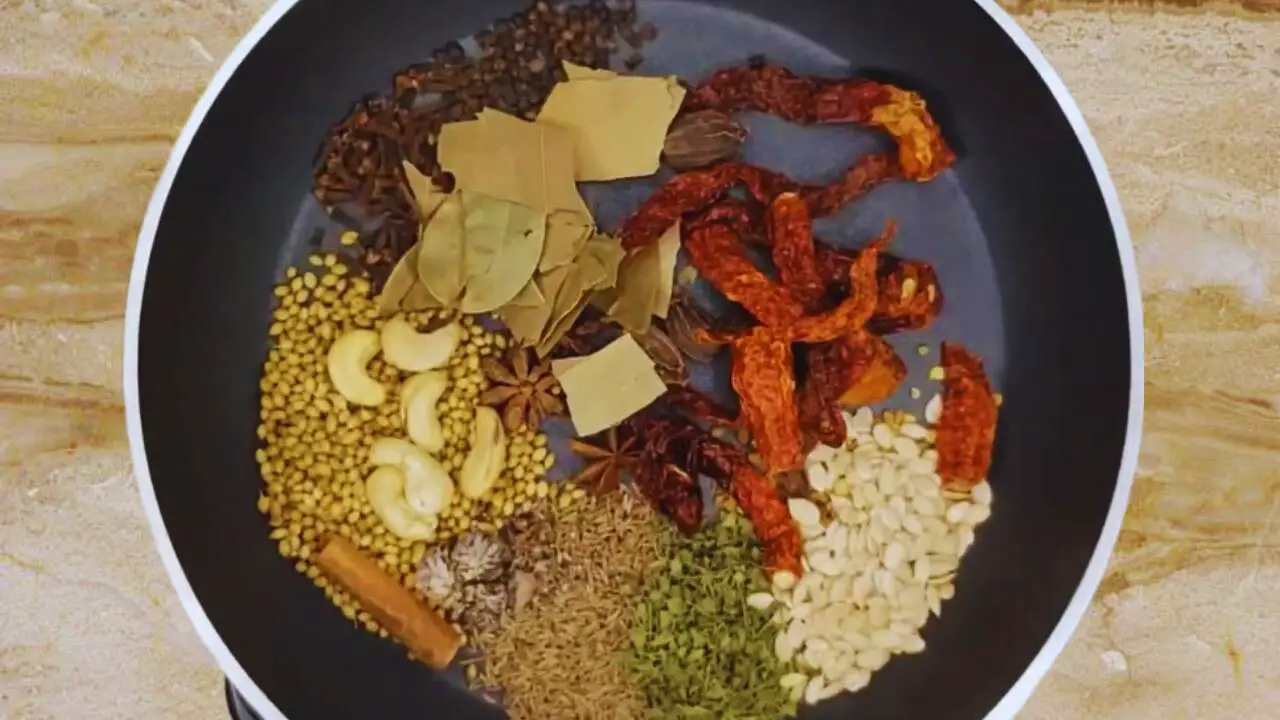 Adding 2 tbsp of coriander seeds, 2 tbsp cumin seeds, 1 tsp fenugreek seeds, 3 pieces of star anise, 1 tsp of cloves, ½ tsp of black pepper, 3 pieces of black cardamom, 8 to 10 pieces of dried Kashmiri red chilies, 3 pieces of bay leaves torn into small pieces, 1 tbsp of dried fenugreek (methi) leaves, 1 small cinnamon stick of about 1 inch in size, 1 piece of nutmeg, 6 pieces of cashew nuts, and 1 tbsp of dried mixed melon seeds (charmagaz) in frying pan