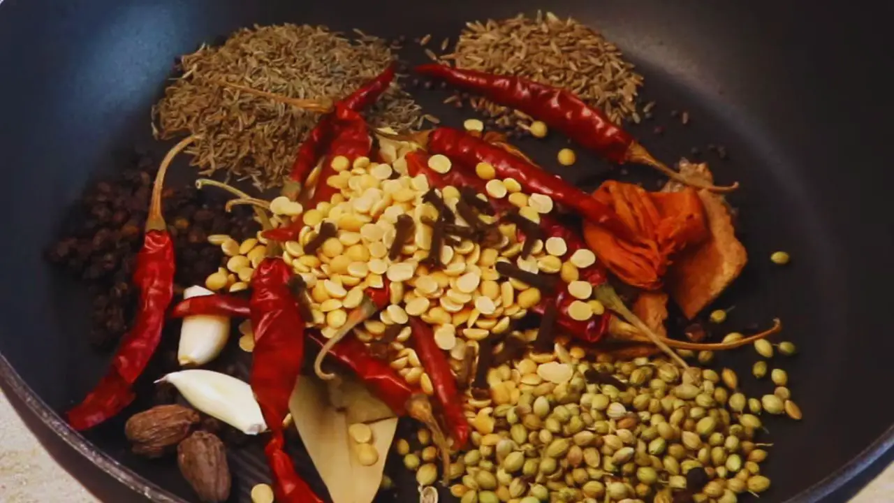 Adding 2 tbsp of coriander seeds, 1.5 tbsp of black pepper, 2 pieces of black cardamom, 1 piece mace, 1 medium piece of star anise, ½ piece of nutmeg, 1 tbsp of cumin seeds, 2 tbsp of caraway seeds (shahi jeera), 2 tbsp of mustard seeds, 2 tbsp of split pigeon pea (arhar dal), 8 pieces of garlic without skin, 2 pieces of medium-sized bay leaves, 12 pieces of dry red chilies, 1 large cinnamon stick (broken into small pieces), 15 pieces of cloves, and 1 piece of dry ginger or about 1.5-inch size into the pan